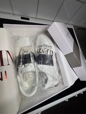 Vita sneakers från Valentino Garavani - Snygga vita sneakers från Valentino Garavani med klassisk låg profil och bred svart logga över sidan. Skorna har vita snören och är tillverkade i slätt läder för en clean och exklusiv look. Perfekta för dig som vill ha en stilren och trendig sneaker. ( använda en gång icke synligt )