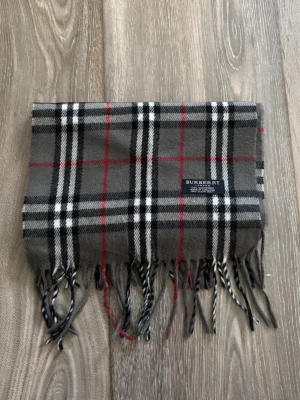 Grå rutig halsduk från Burberry - Klassisk halsduk från Burberry i grått med svart, vitt och rött rutmönster. Tillverkad i 100% kashmir och har fransar i ändarna. Märkt med Burberry London och tillverkad i England. Perfekt accessoar för att lyfta din outfit.