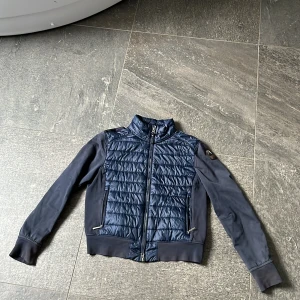 Blå quiltad jacka från Parajumpers - Snygg blå jacka från Parajumpers med quiltad front och ryggparti i blankt material. Jackan har långa ärmar, dragkedja framtill och coolt PJS-märke på ena ärmen. Perfekt för dig som vill ha en sportig och stilren look. Storlek 146-155 liten fläck.