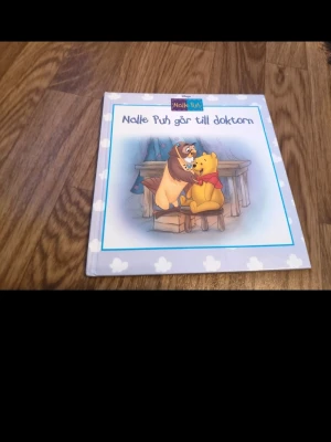 Nalle Puh går till doktorn bok - En söt barnbok med hårda pärmar från Disney om Nalle Puh som besöker doktorn. Omslaget är ljusblått med vita moln och en färgglad illustration av Nalle Puh och Uggla. Perfekt för dig som gillar klassiska sagor och fina illustrationer.
