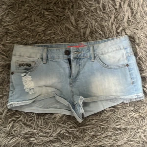 Ljusblå lågmidjade denimshorts med slitningar - Säljer ett par ljusblå lågmidjade denimshorts med slitningar och snygga nitar vid fickan. Klassisk femficksmodell med knappgylf och råa kanter. Perfekta för varma dagar och en avslappnad stil.