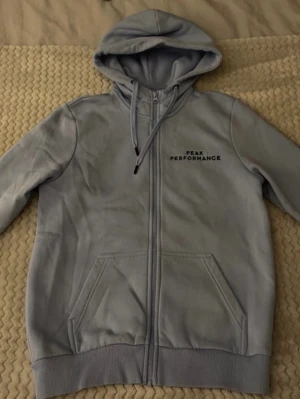 Ljusgrå hoodie från Peak Performance - Snygg ljusgrå hoodie med dragkedja från Peak Performance. Klassisk modell med huva och dragsko, samt fickor framtill. Diskret logga på bröstet. Perfekt för en chill och sportig look.