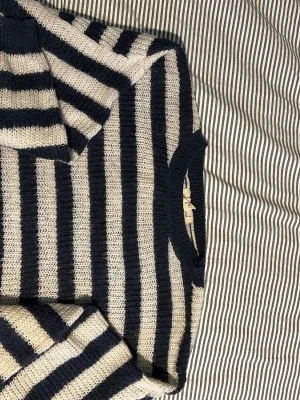 Randig stickad tröja i blått och beige - Stickad tröja med breda horisontella ränder i marinblått och beige. Tröjan har rund halsringning och långa ärmar. Perfekt för en avslappnad och trendig look.