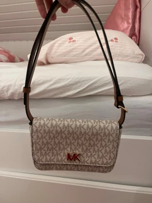 Beige MK handväska  - Snygg beige handväska från Michael Kors med all-over MK-logga och guldfärgad MK-detalj framtill. Väskan har justerbar axelrem i brunt läder och en kompakt, rektangulär form. Perfekt för dig som gillar stilrena accessoarer med tydlig logga.