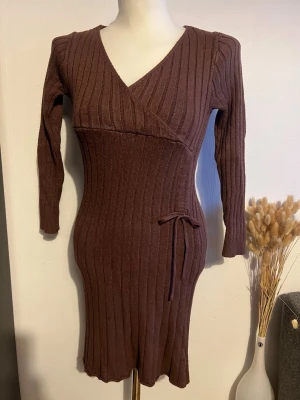 Brun ribbad omlottklänning - Mysig brun ribbad omlottklänning med lång ärm och knyt i midjan. Klänningen har v-ringad hals och är figurnära med stretchigt material, perfekt för en trendig och bekväm look.