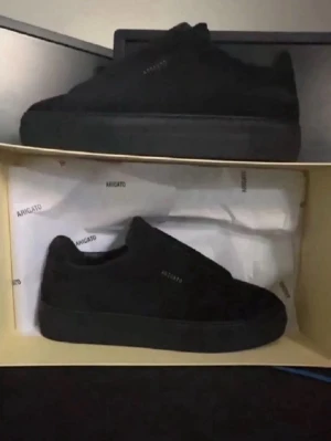 Svarta  Axel Arigato skor  - Stilrena helsvarta sneakers från Axel Arigato i laceless mocka med diskret logga på sidan. Klassisk låg modell med tjock sula och minimalistisk design som passar till det mesta. Perfekta för dig som gillar en clean och modern look.