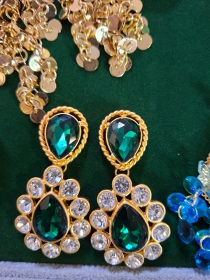 Guldörhängen med gröna stenar - Snygga örhängen i guldton med stora droppformade gröna stenar och klara, runda stenar runtom. Örhängena har en elegant och iögonfallande design med tvinnad guldkant. Perfekta för dig som vill sticka ut med accessoarerna.