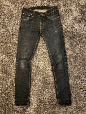 Mörkblå  jeans från Nudie  - Säljer ett par mörkblå  jeans från Nudie Jeans med klassiska kontrastsömmar och fem fickor. Jeansen har en smal passform och är tillverkade i denim med en snygg tvätt. Perfekta för dig som gillar stilrena och tidlösa jeans.
