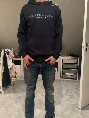Svart hoodie från Lindbergh - Säljer en svart hoodie från Lindbergh med texten 'Copenhagen' tryckt på bröstet. Hoodien har klassisk huva med dragsko och en stor magficka framtill. Perfekt för en avslappnad och stilren look.