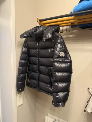 Svart dunjacka från Moncler - Snygg svart dunjacka från Moncler med glansig finish och klassisk patch på ärmen. Jackan har huva, dragkedja framtill och två sidofickor med dragkedja. Perfekt för kalla vinterdagar och även vårdagar, och riktigt trendig look. Den säljs nu när jag växt ut den för ett tag sen! Jackan är 100% äkta och har kvar kvitto. Jackan köptes ny för ca 6700kr