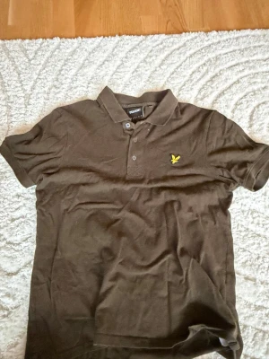 Brun pikétröja från Lyle & Scott - Snygg brun pikétröja från Lyle & Scott med klassisk krage och två knappar framtill. Tröjan har det ikoniska gula logomärket broderat på bröstet och är tillverkad i mjuk bomull. Perfekt till jeans eller shorts för en avslappnad stil. Man får även med en gratis vara på sista bilden🔥🤯