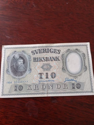Svensk 10-kronorssedel 1954 - Säljer en klassisk svensk 10-kronorssedel från 1954, utgiven av Sveriges Riksbank. Sedeln har porträtt av Gustav Vasa på framsidan och riksvapnet med tre kronor på baksidan. Färgerna är främst grå, svart och vit med detaljerade mönster.