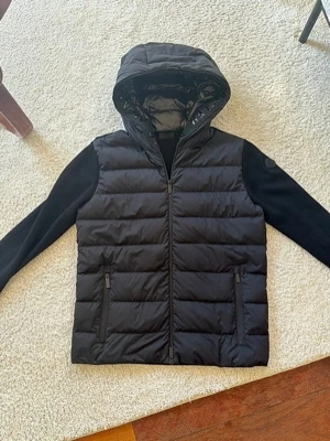 Moncler Stickad Jacka Med Huva  - Snygg svart pufferjacka med vadderad framsida och huva. Ärmar och rygg är i mjukt stickat material för extra komfort. Jackan har två dragkedjeförsedda fickor framtill och stängs med dragkedja. Perfekt för kyliga dagar och en stilren look.