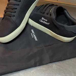 Svarta Saint Laurent sneakers - Snygga svarta sneakers från Saint Laurent med vit sula och broderad logga på sidan. Skorna har klassisk snörning, rund tå och är tillverkade i canvas och skinn. Perfekta för dig som vill ha en stilren och exklusiv look med streetkänsla.