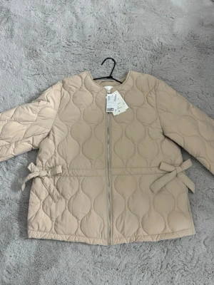 Beige quiltad jacka  - Supersnygg beige pufferjacka med quiltat mönster och dragkedja framtill. Jackan har rund halsringning och dekorativa rosetter i midjan på båda sidor. Perfekt för kalla dagar och ger en trendig look. Materialet är mjukt och värmande.