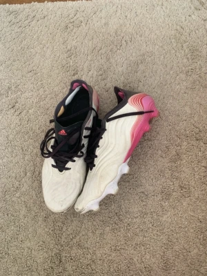 Adidas copa sense elite  - Säljer ett par Adidas copa sense fotbollsskor i vitt med svarta detaljer och rosa gradient på hälen. Skorna har snörning, vågigt mönster på ovandelen och robusta dobbar för bra grepp på planen. Perfekta för dig som vill sticka ut på matchen!