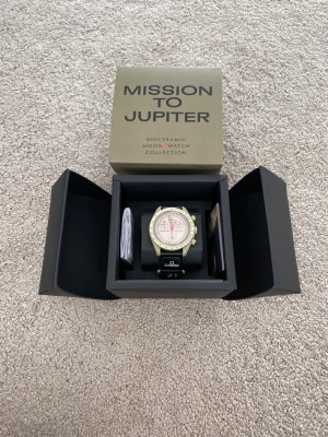 Omega x Swatch Mission to Jupiter - Säljer en Omega x Swatch Speedmaster MoonSwatch Mission to Jupiter. Klockan har en beige boett i biokeramik, vit urtavla med orange detaljer och svart textilband med Omega-logga. Kommer i originalbox med tillbehör. Perfekt för dig som gillar unika klockor!