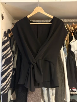 Zara topp - Stilren svart omlottopp med djup v-ringning och knyt i midjan. Toppen har breda axelband och är ärmlös, vilket ger en elegant och modern look. Perfekt att matcha med både jeans och kjol för en trendig outfit.