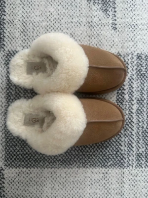 Uggs Bruna/ Chesnut tofflor från UGG - Mysiga tofflor från UGG i brun/ chestnut färg med fluffig vit ull i insidan. Klassisk slip-in modell med rund tå och mjuk innersula i ull. Perfekta för att hålla fötterna varma hemma. UGG-logga på sidan.