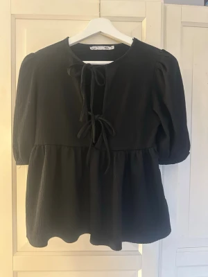 Svart blus med puffärm och knyt - Svart blus från lager 157 med puffiga trekvartsärmar och två dekorativa knytband framtill. Tillverkad i ett mjukt, strukturerat tyg som ger en avslappnad men ändå trendig look. Perfekt att styla med jeans eller kjol för en cool vibe.