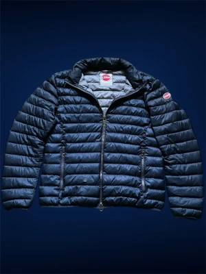 Mörkblå dunjacka från Colmar - Snygg mörkblå dunjacka från Colmar med quiltad design och två dragkedjeförsedda fickor framtill. Jackan har hög krage och Colmar-logga på ärmen. Perfekt för kalla vinterdagar och riktigt lätt att bära. Är ett litet hål som det finns bild på men där räcker det med att sätta på en tyg lapp över.