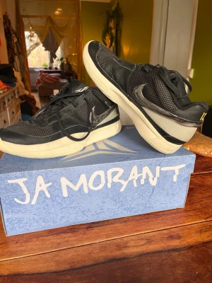 Nike Ja Morant svarta sneakers - Snygga Ja Morant sneakers från Nike i svart mesh och syntet med vita och beige detaljer. Lågt skaft, gula detaljer på hälen och coola loggor. Perfekta för dig som vill sticka ut med stilrena och sportiga skor.