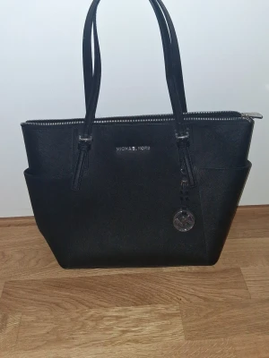 Svart handväska från Michael Kors - Stilren svart handväska från Michael Kors i tåligt saffianoläder. Väskan har två långa handtag, silverfärgade detaljer och ett hängande MK-emblem. Praktisk dragkedja upptill och sidofickor för extra förvaring. Perfekt för dig som gillar klassisk och trendig design. (Pris kan diskuteras)