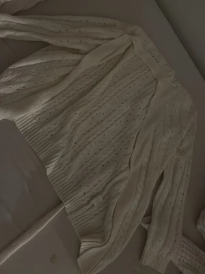 Vit kabelstickad kofta med knytband - Säljer en krämvit kabelstickad kofta med dekorativa hålmönster och breda ribbade muddar. Den är från Shein och har använts ett par gånger mer är fortfarande i bra skick. Den är i strl S men kan passa dig i strl Xs också. 