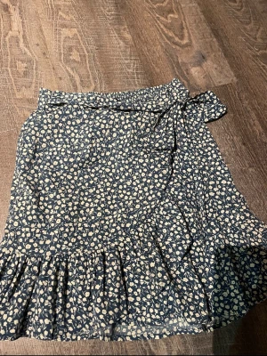 Blommig omlottkjol från ONLY - Supersöt blå omlottkjol från ONLY med vitt småblommigt mönster och volangkant. Kjolen har knytband i midjan och är gjord i ett lätt, flowy material som känns perfekt för vår och sommar. Passar dig som gillar en feminin och trendig stil.