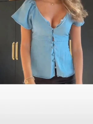 Blå v-ringad blus med puffärm - Snygg blå blus med v-ringning och puffärmar. Blusen har knappar framtill och är lätt figursydd för en feminin siluett. Perfekt till jeans eller kjol för en trendig look.