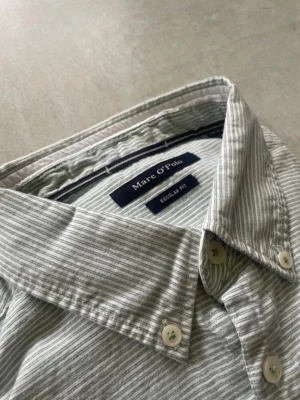 Randig kortärmad skjorta Marc O'Polo - Snygg ljusgrå och vit randig skjorta från Marc O'Polo med korta ärmar och klassisk krage. Skjortan har bröstficka och knappar framtill. Regular fit och tillverkad i mjuk bomull, perfekt för en avslappnad stil.