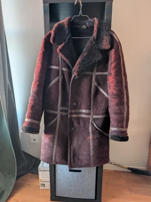 Vinröd fårskinnskappa med teddyfoder - Säljer en lång vinröd fårskinnskappa med mysigt svart teddyfoder och stora slag. Kappan har markerade sömmar, knäppning med stora knappar och två snedställda fickor framtill. Perfekt för kalla dagar och ger en cool vintagekänsla.