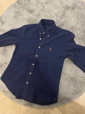 Mörkblå skjorta från Ralph Lauren - Klassisk mörkblå skjorta från Ralph Lauren med broderad logga på bröstet. Skjortan har knappar framtill, button-down krage och långa ärmar. Tillverkad i mjuk bomull som känns skön mot huden. Perfekt för en stilren och avslappnad look.