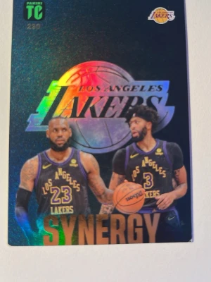 Los Angeles Lakers samlarkort Synergy - Unikt samlarkort från Panini med Los Angeles Lakers-tema. Kortet har holografisk yta och motiv av två Lakers-spelare i matchställ, samt texten 'Synergy' i guld. Perfekt för basketfans och samlare.