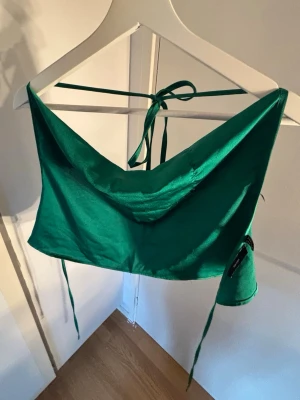 Grön satintopp med knyt - Snygg grön satintopp med tunna band som knyts i nacken och ryggen. Toppen har en draperad framsida och croppad passform, perfekt för att styla med höga jeans eller kjol. Materialet har en glansig finish som ger en lyxig känsla.