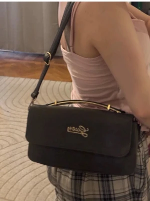 Svart axelväska från Guess - Snygg svart axelväska från Guess med guldfärgade detaljer och logga framtill. Väskan har en stilren, rektangulär form och justerbar axelrem. Perfekt för dig som vill ha en trendig och praktisk väska till vardagen.