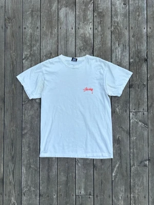 Vit Stüssy t-shirt med tryck - Vit t-shirt från Stüssy med röd logga på bröstet och stort grafiskt tryck på ryggen där en person målar Stüssy-loggan. Klassisk passform och rund halsringning. Perfekt för dig som gillar streetwear och vill sticka ut med en ikonisk design.