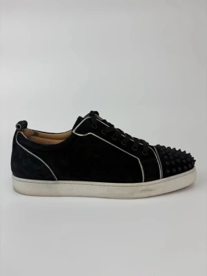 Svarta sneakers med nitar från Louboutin - Svarta sneakers i mocka från Christian Louboutin med vita kontrastsömmar och coola svarta nitar på tån. Skorna har vita sulor och svarta skosnören, samt en unik design som sticker ut. Perfekta för dig som vill ha något extra till din outfit.