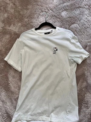 Vit Snoopy t-shirt från H&M - Vit t-shirt från H&M med Snoopy-tryck på bröstet och stort färgglatt graffitiinspirerat tryck med Snoopy och Woodstock på ryggen. Klassisk passform och rund halsringning. Perfekt för dig som gillar streetstyle och tecknade motiv.