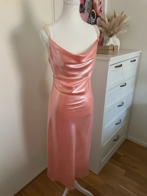 Glansig rosa satinklänning med pärlband - Snygg rosa satinklänning med glansig finish och pärlprydda axelband. Klänningen har en draperad urringning och är ärmlös med en figurnära passform. Perfekt för dig som vill sticka ut med en elegant och trendig look.