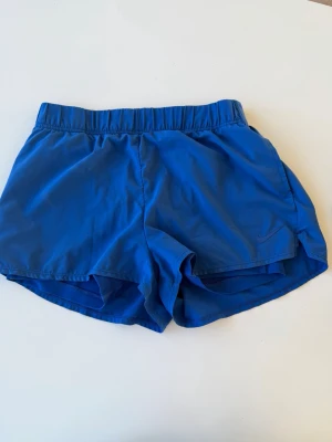 Blå träningsshorts från Nike - Snygga blå träningsshorts från Nike med elastisk midja och klassisk sportig look. Tillverkade i lätt och snabbtorkande syntetmaterial, perfekta för löpning eller gym. Diskret Nike-logga framtill och har undershorts. 