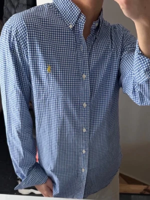 Blårutig skjorta från Ralph Lauren - Snygg blårutig skjorta från Ralph Lauren i slim fit-modell. Skjortan är tillverkad i stretchig bomull och har klassisk krage, knappar framtill och den ikoniska gula loggan broderad på bröstet.perfekt till sommaren 
