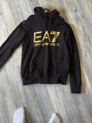 Svart EA7 Emporio Armani hoodie - Snygg svart hoodie från EA7 Emporio Armani med stor guldfärgad logga på bröstet. Klassisk huva med dragsko och magficka framtill. Tillverkad i mjukt material som känns skönt mot huden. Perfekt för en trendig och avslappnad stil.