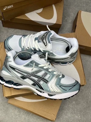 ASICS vita och silver sneakers - Snygga ASICS sneakers i vitt och silver med svarta detaljer. Ovandelen är i mesh och syntetmaterial för bra andningsförmåga och komfort. Klassisk ASICS-logga på sidan och vadderad plös. Perfekta för en sportig och trendig look.