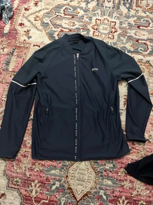 Svart sportjacka från BOSS - Snygg svart sportjacka från BOSS med hel dragkedja och diskreta vita detaljer. Jackan har tunna ränder på axlar och rygg, två fickor med dragkedja och logga på bröstet. Perfekt för en stilren och sportig look.
