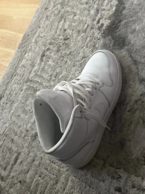 Vita Nike high top sneakers - Säljer ett par helvita high top sneakers från Nike. Skorna har klassisk design med snörning, diskret Nike-logga på sidan och är tillverkade i syntetmaterial och textil. Perfekta för dig som gillar stilrena och sportiga sneakers.