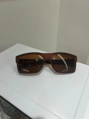 Bruna fyrkantiga solglasögon från Prada - Snygga solglasögon från Prada med fyrkantig form och bruna bågar samt bruna glas. Diskret Prada-logga på sidan. Perfekt accessoar för att lyfta din stil med en modern och trendig look.