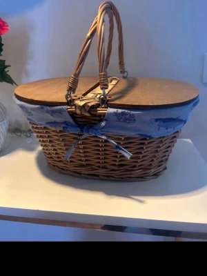Klassisk picknickkorg i rotting - Charmig picknickkorg i flätad rotting med lock i trä och dubbla handtag. Korgen är dekorerad med ett blått tygband med fiskmotiv och rosett framtill. Perfekt för utflykter eller som snygg förvaring hemma.