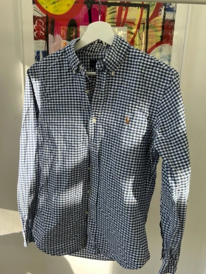 Blårutig skjorta från Ralph Lauren - Snygg blårutig skjorta från Ralph Lauren i slim fit-modell. Skjortan har klassisk krage, knappar framtill och en liten broderad logga på bröstet. Perfekt för dig som gillar stilrena och tidlösa plagg.