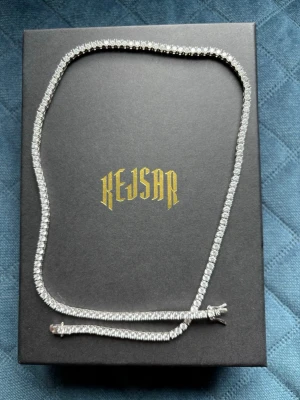 Silverfärgat tennis-halsband Kejsar 3mm nyskick  - Stilrent tennis-halsband från Kejsar i silverfärg med gnistrande stenar längs hela kedjan. Halsbandet har en klassisk, rak form och ger en lyxig känsla. Perfekt för dig som vill sticka ut med en glittrig accessoar. Använd max 5 gånger sö är fortfarande i nyskick.